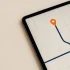 Minimal overhead view of a tablet showing a simple route map with an orange location pin symbolizing flatbed trucking route planning and permits
